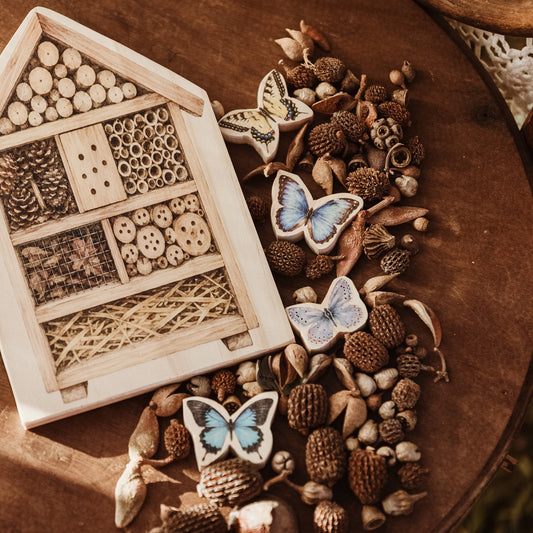 A wooden house-shaped insect hotel with multiple compartments filled with natural materials, arranged on a rustic wooden surface alongside pinecones, acorns, dried leaves, and three ceramic butterflies.