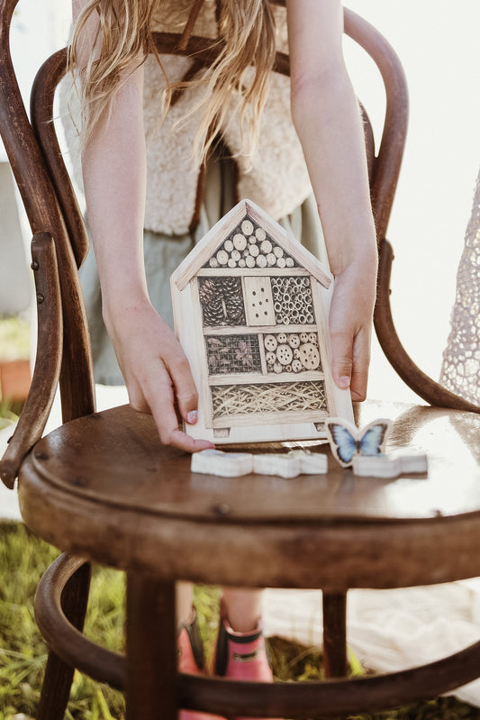 A child holds a small wooden house-shaped insect hotel with patterned panels, seated on a rustic wooden chair outdoors.