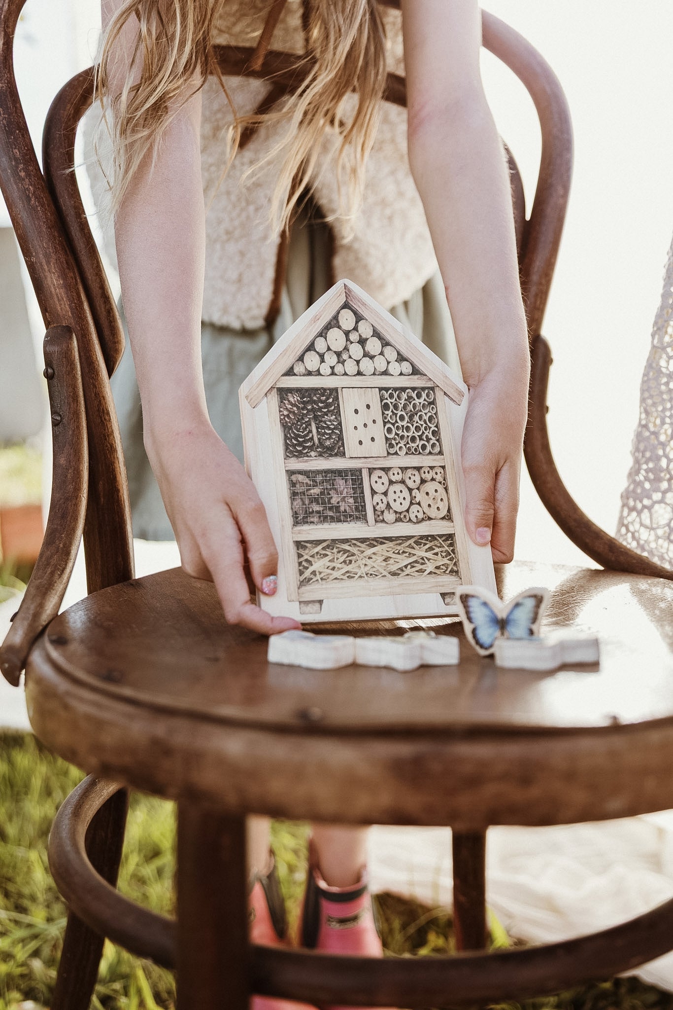 A child holds a small wooden house-shaped insect hotel with patterned panels, seated on a rustic wooden chair outdoors.